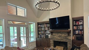 A spacious living room with a large flat-screen TV, a stone fireplace, and a ceiling light fixture.