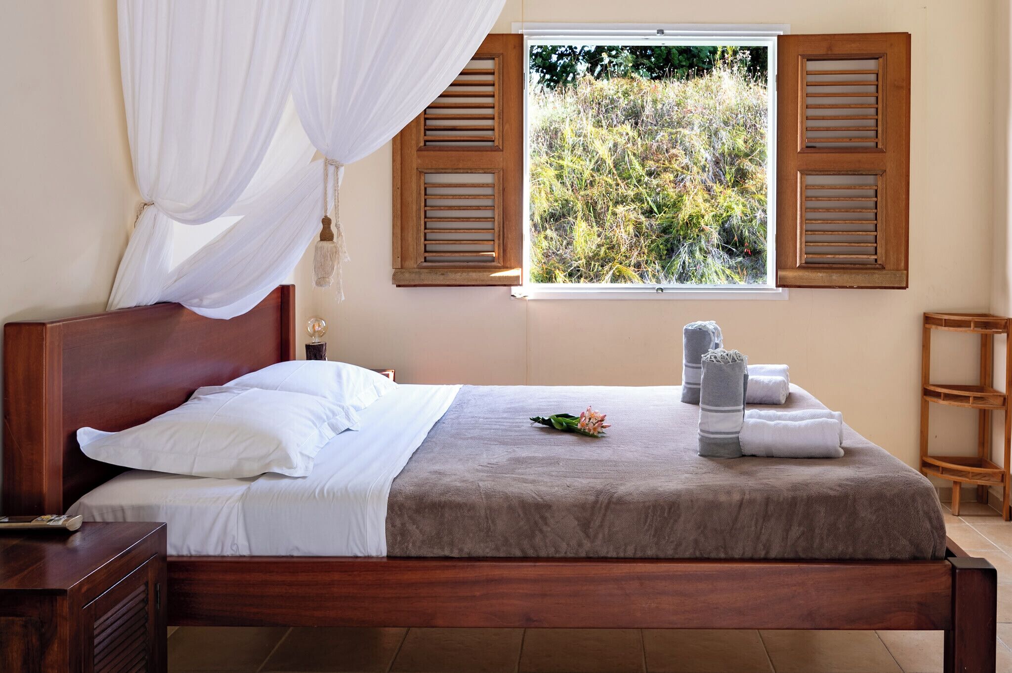Cama bem arrumada com lençóis brancos, um espalha-manto marrom e um buquê de flores. Há persianas de madeira na janela e uma mesa de cabeceira de madeira com uma luminária ao lado da cama.