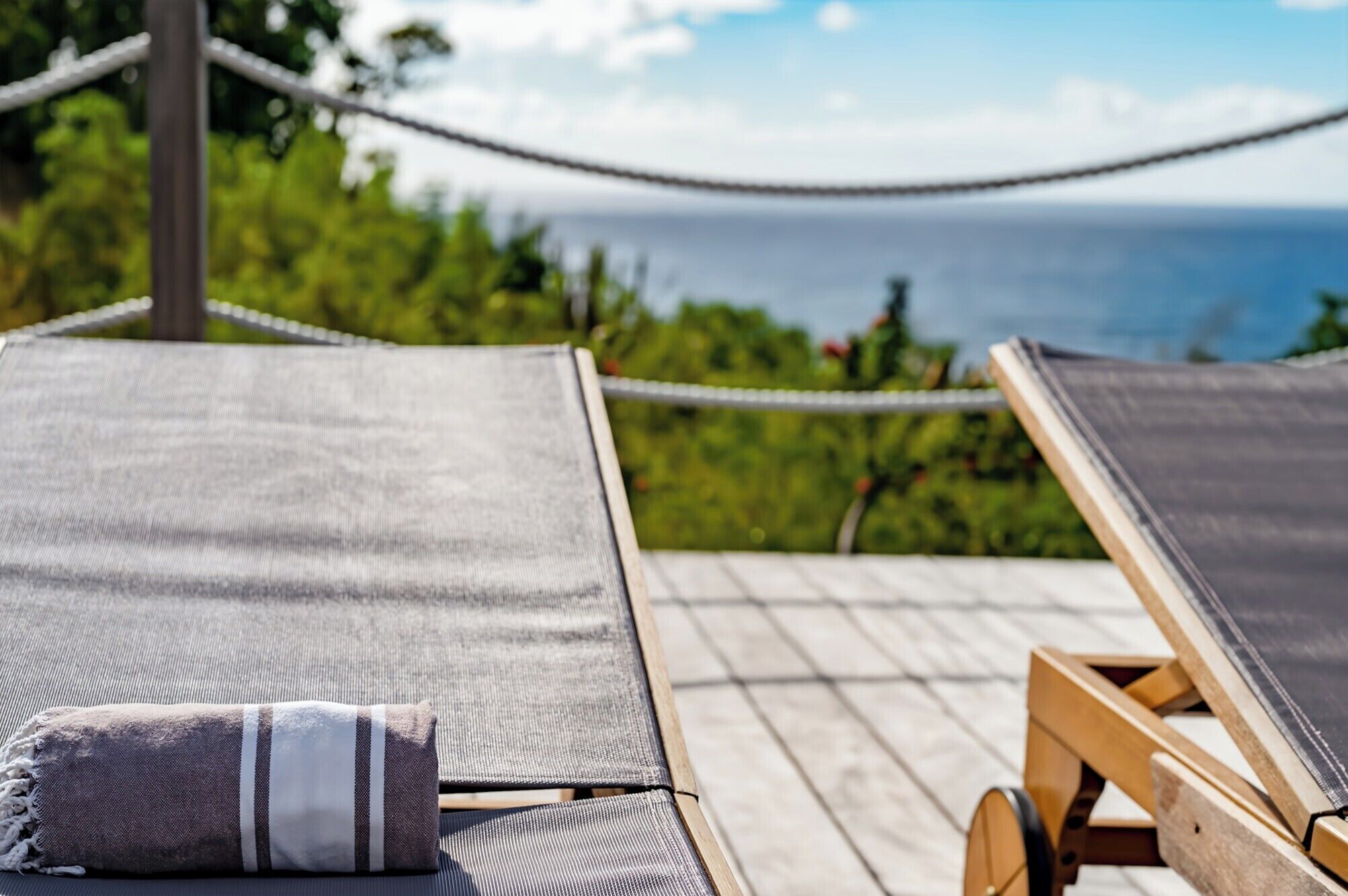 Una terraza de madera con dos sillones de descanso, uno con cojín y el otro vacío. Una hamaca está colgada entre las sillas, y se ve el océano al fondo.
