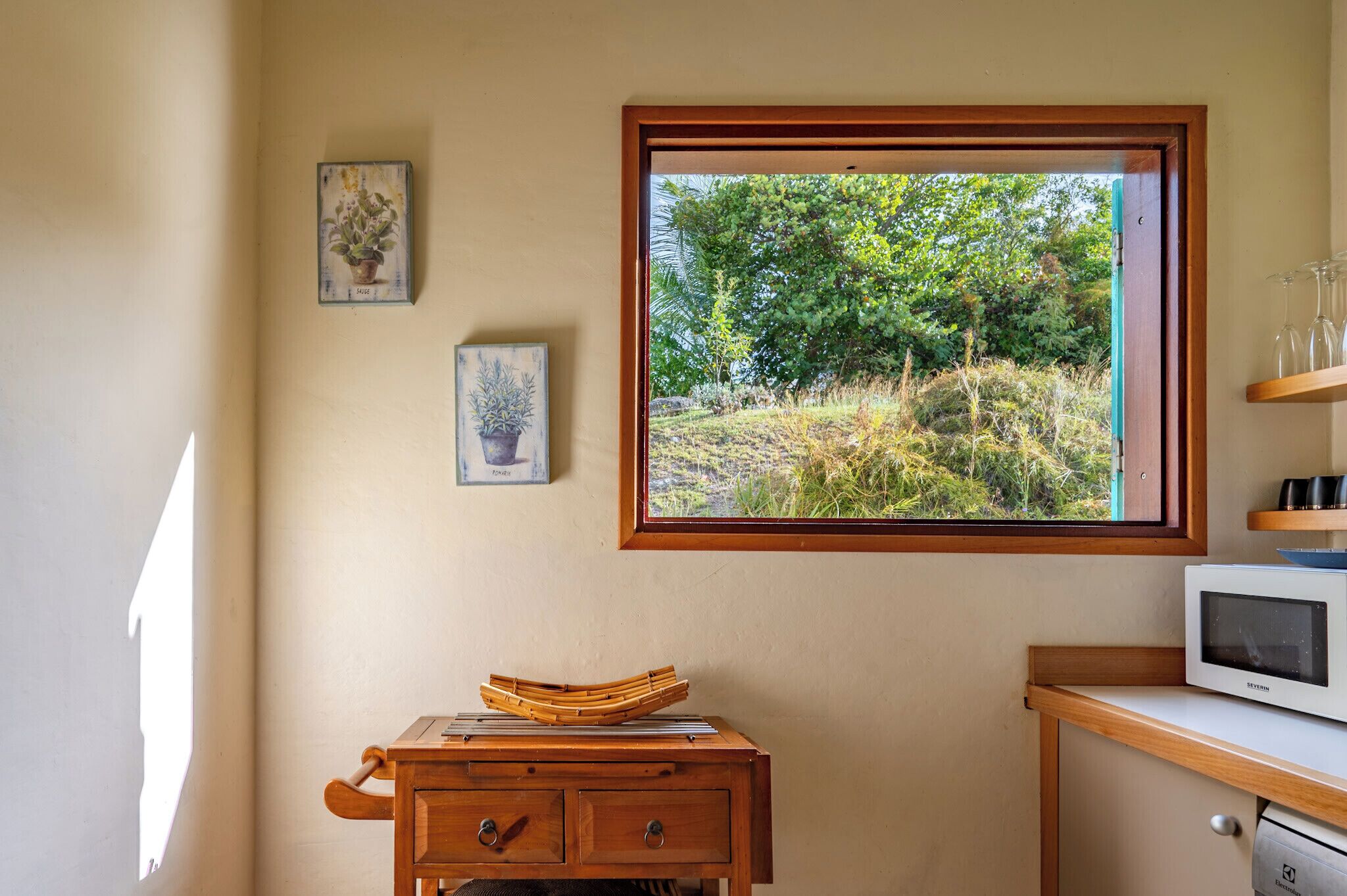 A room with a wooden desk, a small cabinet, a microwave, and a window with a view of greenery.