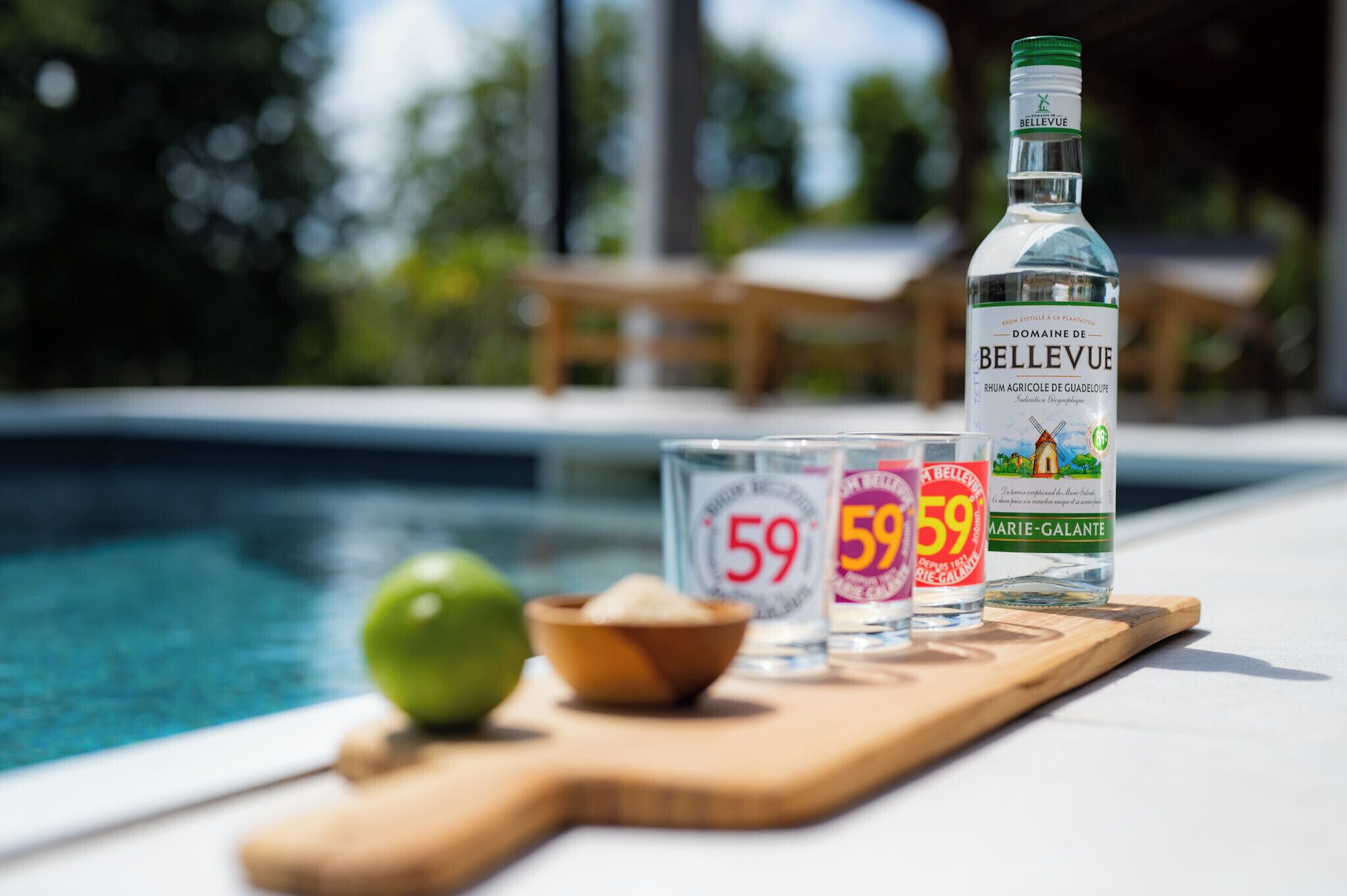 A bottle of Bellevue rum, a lime, and two glasses with the number 59 on a wooden board by a pool.