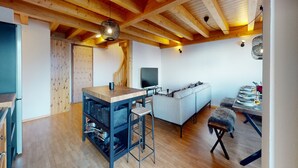 Una habitació àmplia amb sostre de fusta, un sofà, taula de menjador preparada per a quatre i zona de cuina amb un tamboret.