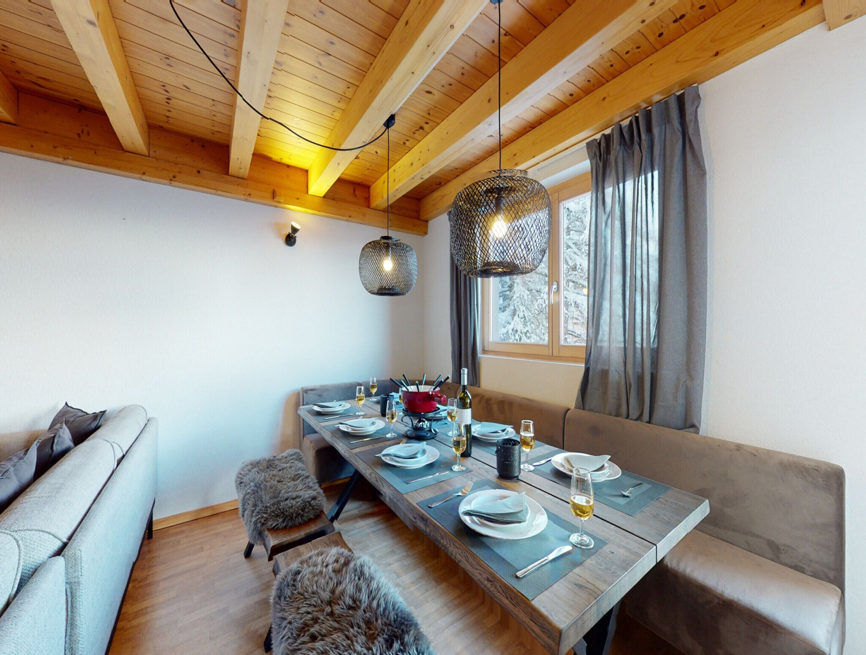 Un comedor con mesa de madera dispuesta para cuatro personas, un sofá y una ventana con cortinas.