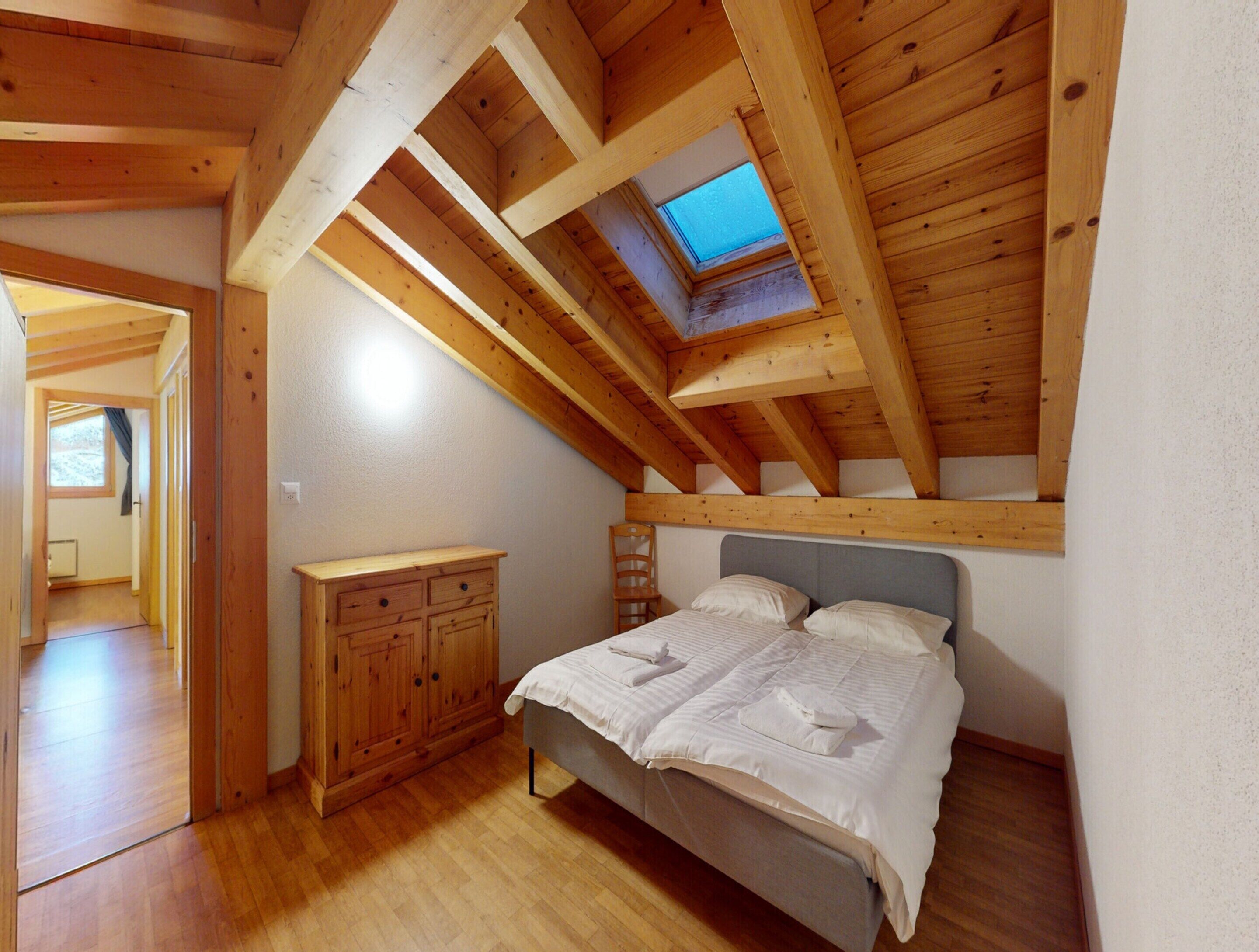 Ein Schlafzimmer mit schräg laufender Decke, einem Dachfenster, einem Bett mit weißer Bettwäsche, einem Holzschrank und einem kleinen Stuhl.