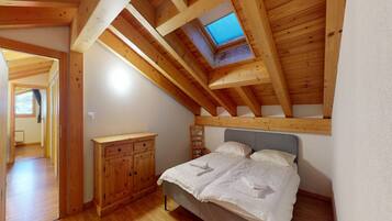 Een slaapkamer met een schuine kap, dakraam, een bed met wit beddengoed, een houten dressoir en een kleine stoel.