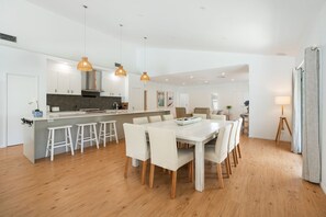 A modern kitchen with white cabinets, a central island with stools, pendant lighting, and a dining area with a white table and chairs.