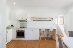 A modern kitchen with white cabinetry, a built-in oven, a stove, and two bar stools. - Winders Beach House (Marcoola)
