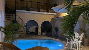 Uma área de piscina externa com espreguiçadeira de madeira, cadeiras brancas e plantas em vasos.