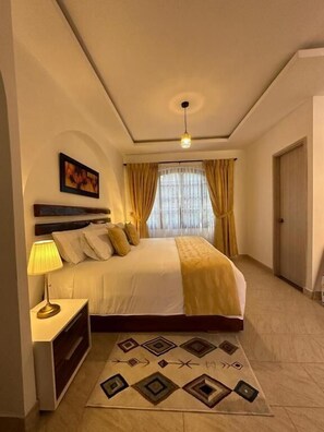 Un dormitorio con cama, mesita de noche, lámpara, luz de techo, puerta y ventana con cortinas.
