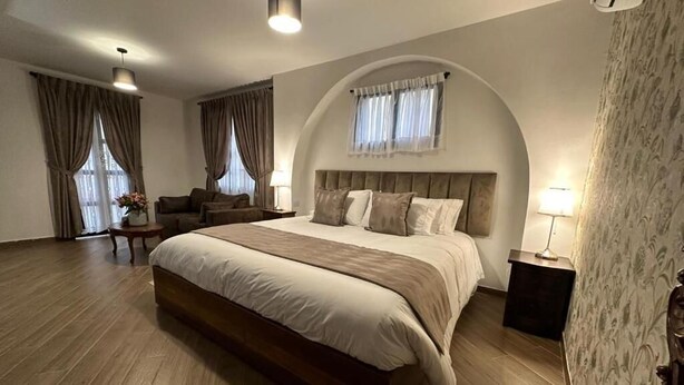 Um quarto espaçoso com uma cama grande, um espaço de estar com uma poltrona e uma mesa, e uma janela com cortinas.