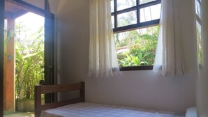 Une petite chambre avec un lit simple, une tête de lit en bois, une fenêtre avec des rideaux et une vue sur la verdure.