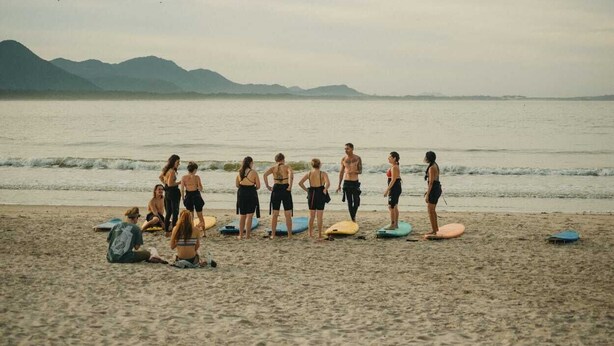 Une plage où l’on voit des gens debout et assis, dont certains tiennent des planches de surf, avec des montagnes en arrière-plan.