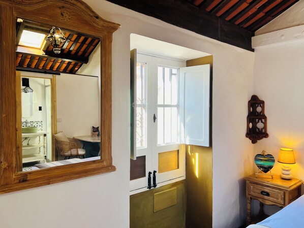 Una habitación con techo de madera, un espejo, una ventana con puerta abierta, una mesita de noche con lámpara y una estantería en la pared.