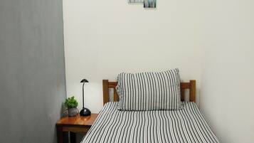 Una cama individual con una colcha a rayas y almohadas, una mesita de noche con una lámpara y una planta pequeña, y una pared con dos cuadros abstractos.