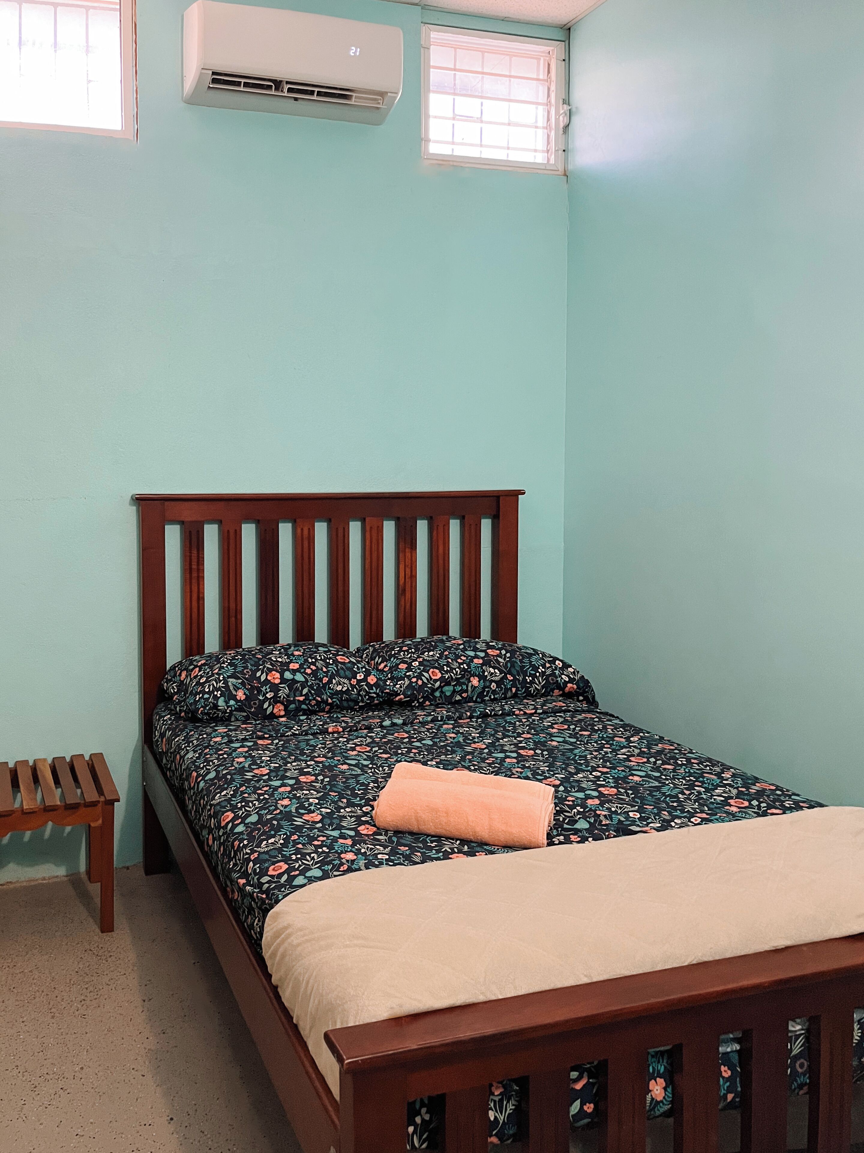 A bedroom with a wooden bed, a small wooden stool, a window, and an air conditioning unit.