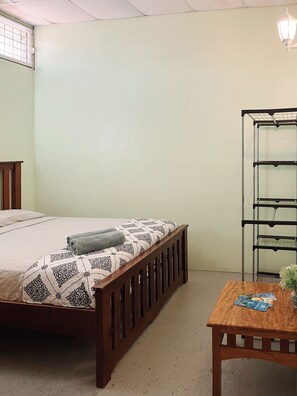 Quarto com cama, mesa de cabeceira de madeira, uma pequena mesa com um vaso e uma estante de metal.
