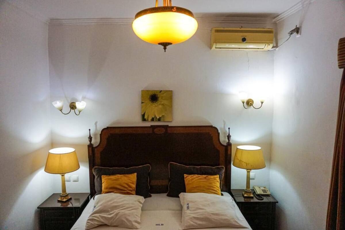A hotel room with a wooden headboard, two bedside lamps, a single bed with white and yellow bedding, a wall-mounted air conditioner, and a painting of a sunflower.