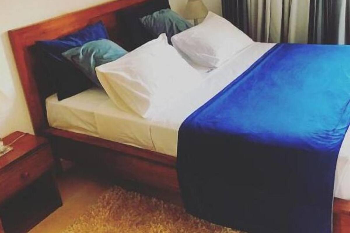 A neatly made bed with a blue blanket and white sheets, a wooden headboard, and a nightstand with a lamp.