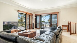 A spacious living room with a large sectional sofa, a coffee table, and a flat-screen TV. There are large sliding glass doors leading to an outdoor area with a view of trees and a mountain. - Beach Front Beach Break - Entire Home (Byron Bay)