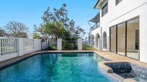 A swimming pool with a tiled edge, surrounded by a white fence, and a building with large windows and a brick facade. - Beach Front Beach Break - Entire Home (Byron Bay)