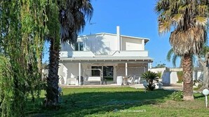 Ein modernes, zweistöckiges Haus mit überdachter Veranda, umgeben von üppiger Vegetation und Palmen.