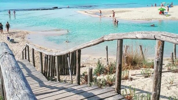 Eine Holzpromenade führt zu einem Strand mit klarem türkisblauem Wasser und Personen in der Ferne.