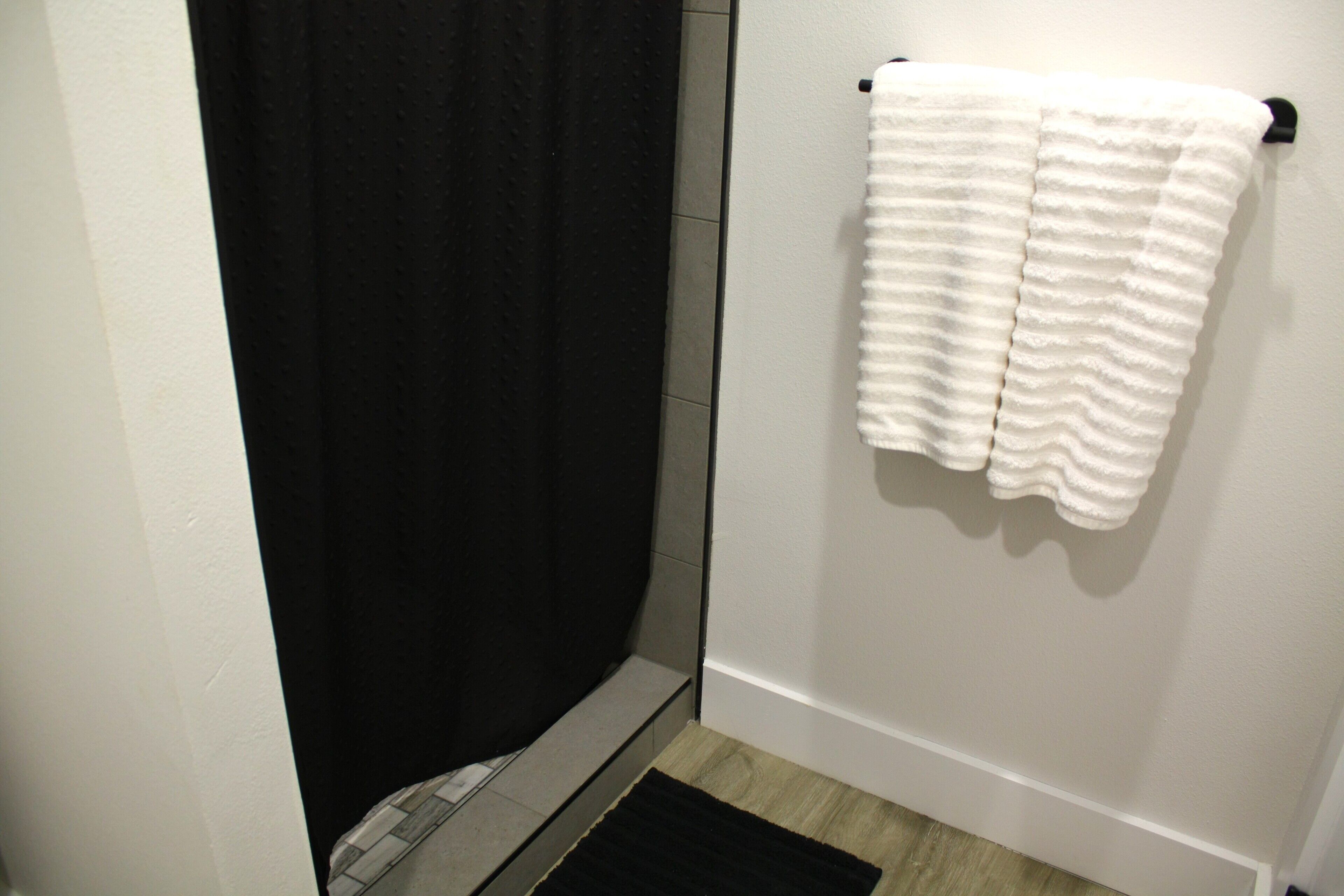 A bathroom with a black shower curtain, a white towel hanging on a rack, and a black rug on the floor.