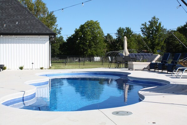 A swimming pool with a curved edge, surrounded by a white deck. There are lounge chairs and a covered area with a table and chairs. - Modern Family Getaway! Heated Pool, Hot tub Theatre Room, Game room! (Joplin)
