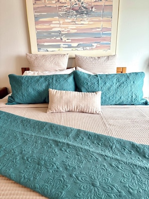 Una cama con colcha color turquesa y almohadas, una colcha blanca y un cuadro en la pared.