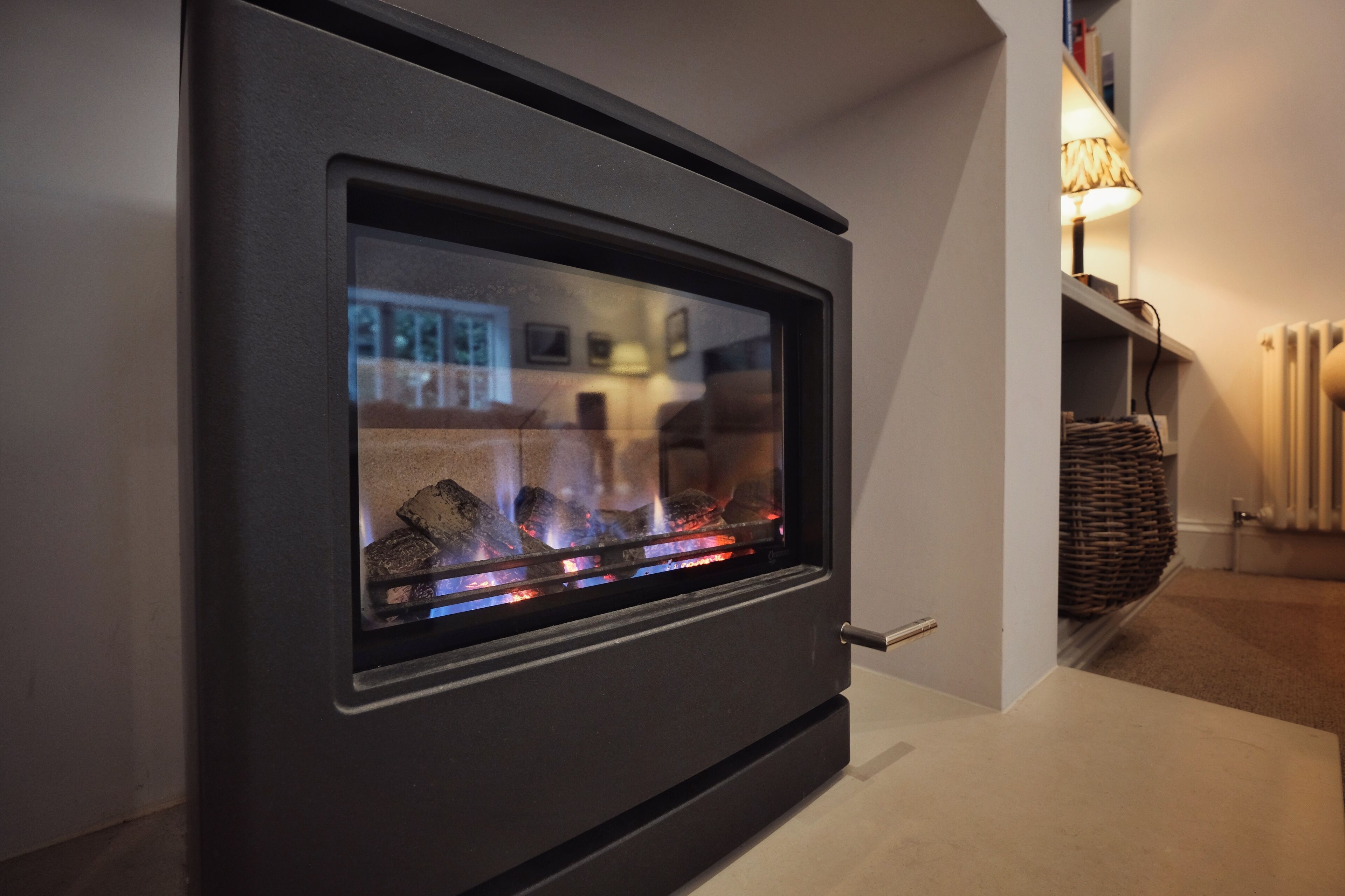 A modern fireplace with a glass front, a white wall, and a bookshelf with a lamp.