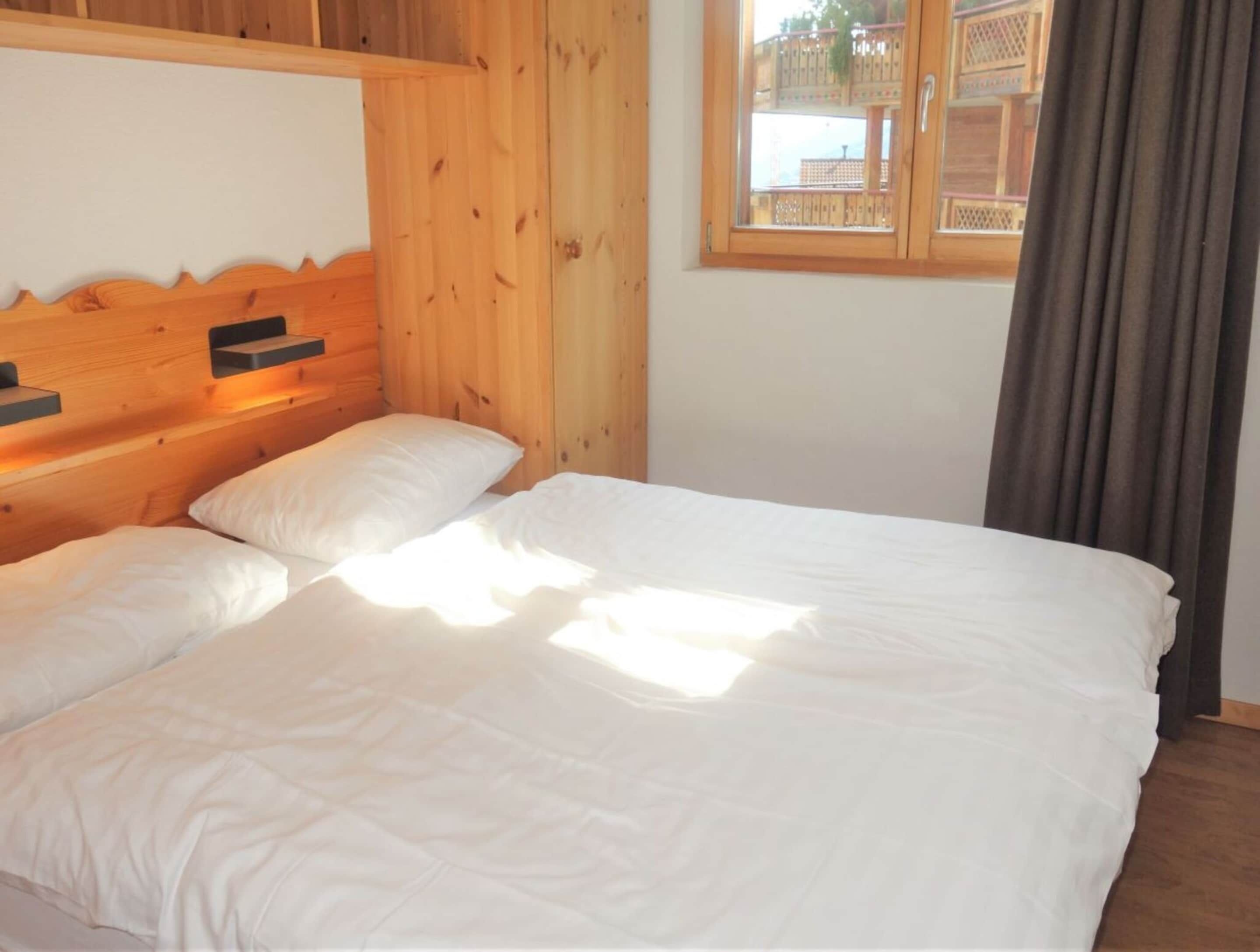A wooden cabin-style room with a bed, a window with a view of a balcony, and a curtain.