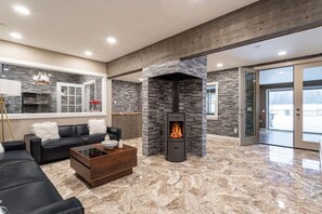 A modern living room with a stone fireplace, a black leather sofa, a coffee table, and a large window. - Equestrian Estate | Indoor Pool, Theater, Game Rm (Schoharie)