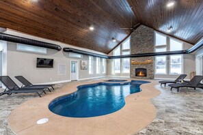 An indoor swimming pool with a stone fireplace, lounge chairs, and large windows.