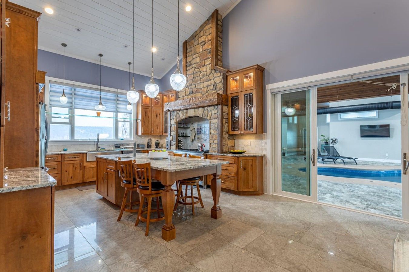 A spacious kitchen with a large island, stone fireplace, and a view of a swimming pool through glass doors.