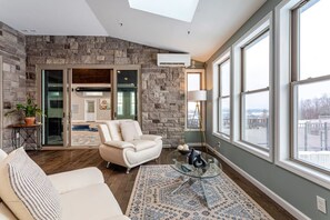 A modern living room with a stone wall, a glass door leading outside, a white sofa, a glass coffee table, and a large window with a view. - Equestrian Estate | Indoor Pool, Theater, Game Rm (Schoharie)