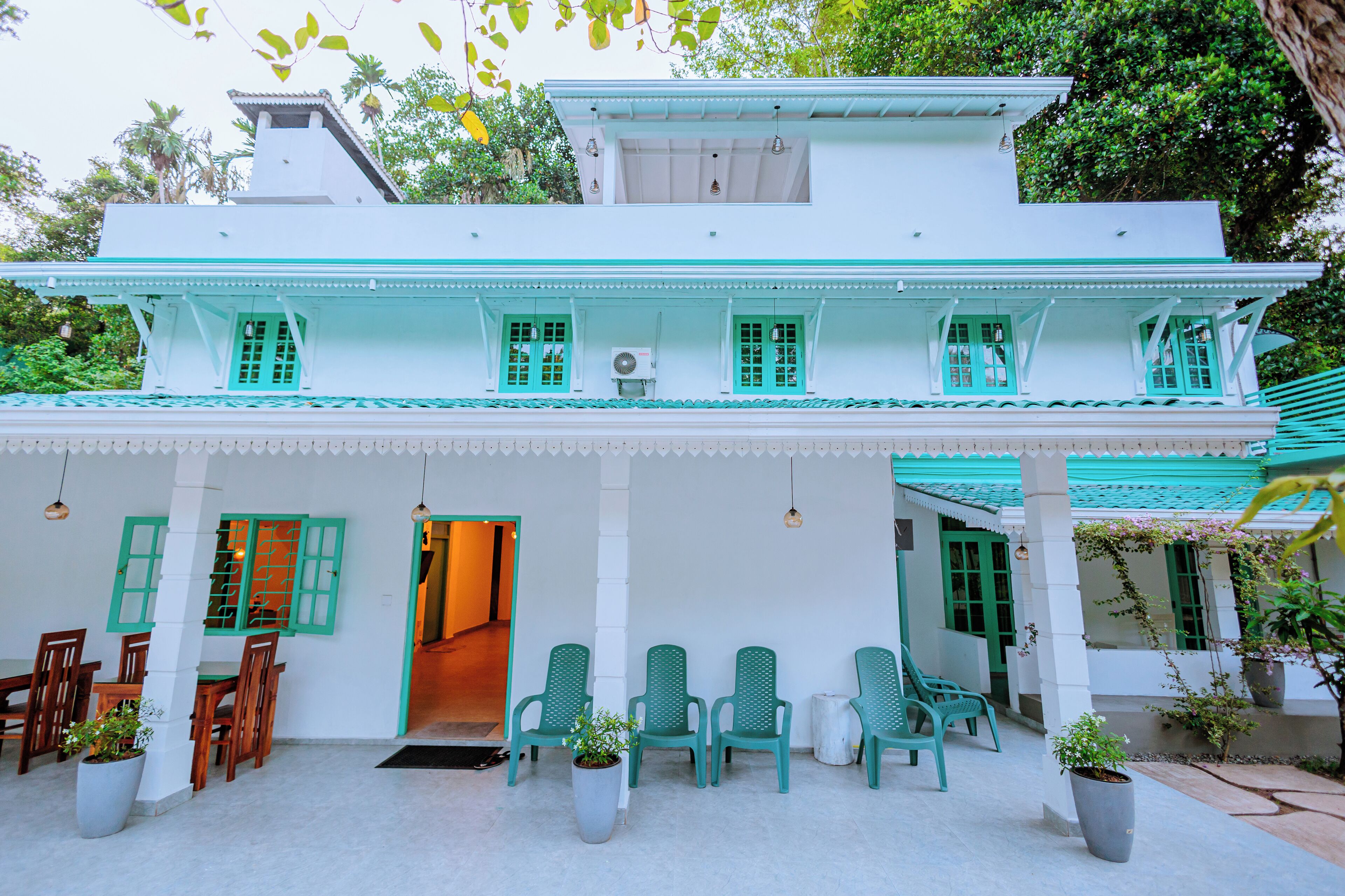 A two-story building with a white exterior and green accents, featuring a covered patio area with chairs and potted plants.