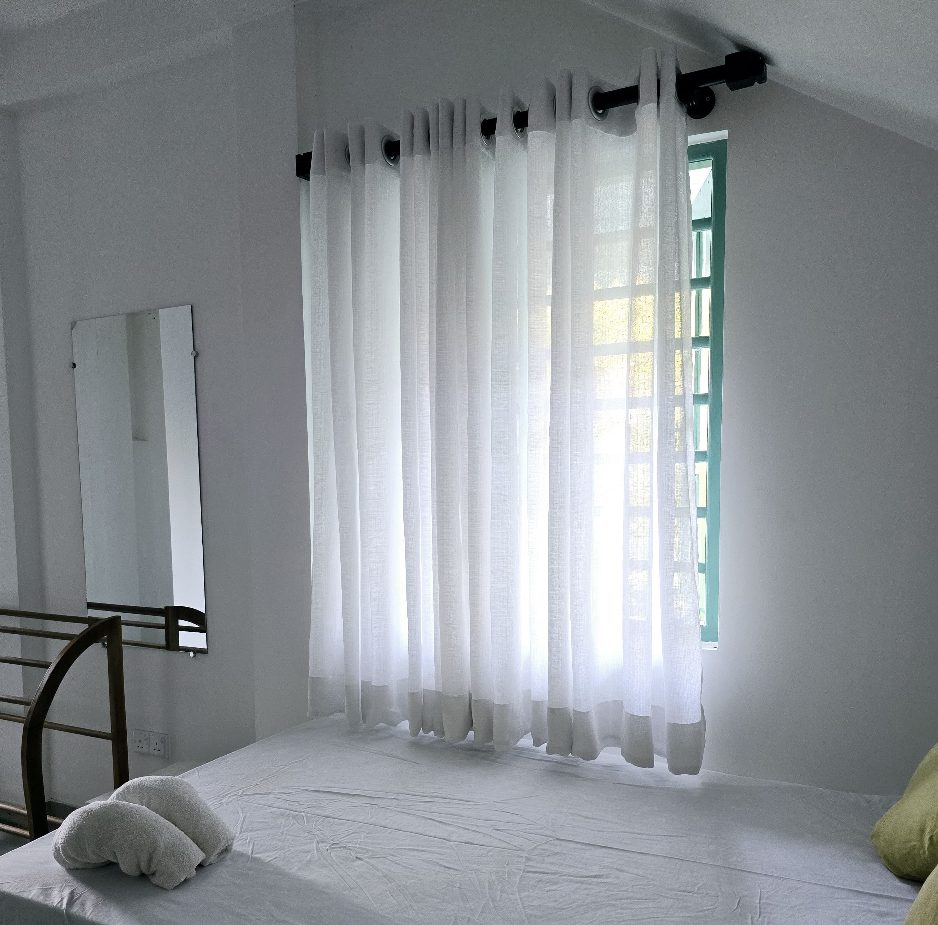 A room with a bed, a window with sheer curtains, a mirror on the wall, and a green window frame.