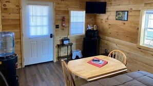 A wooden cabin interior with a dining table, chairs, a water cooler, a fire extinguisher, and a television mounted on the wall. - Canal Cabins (Paw Paw)