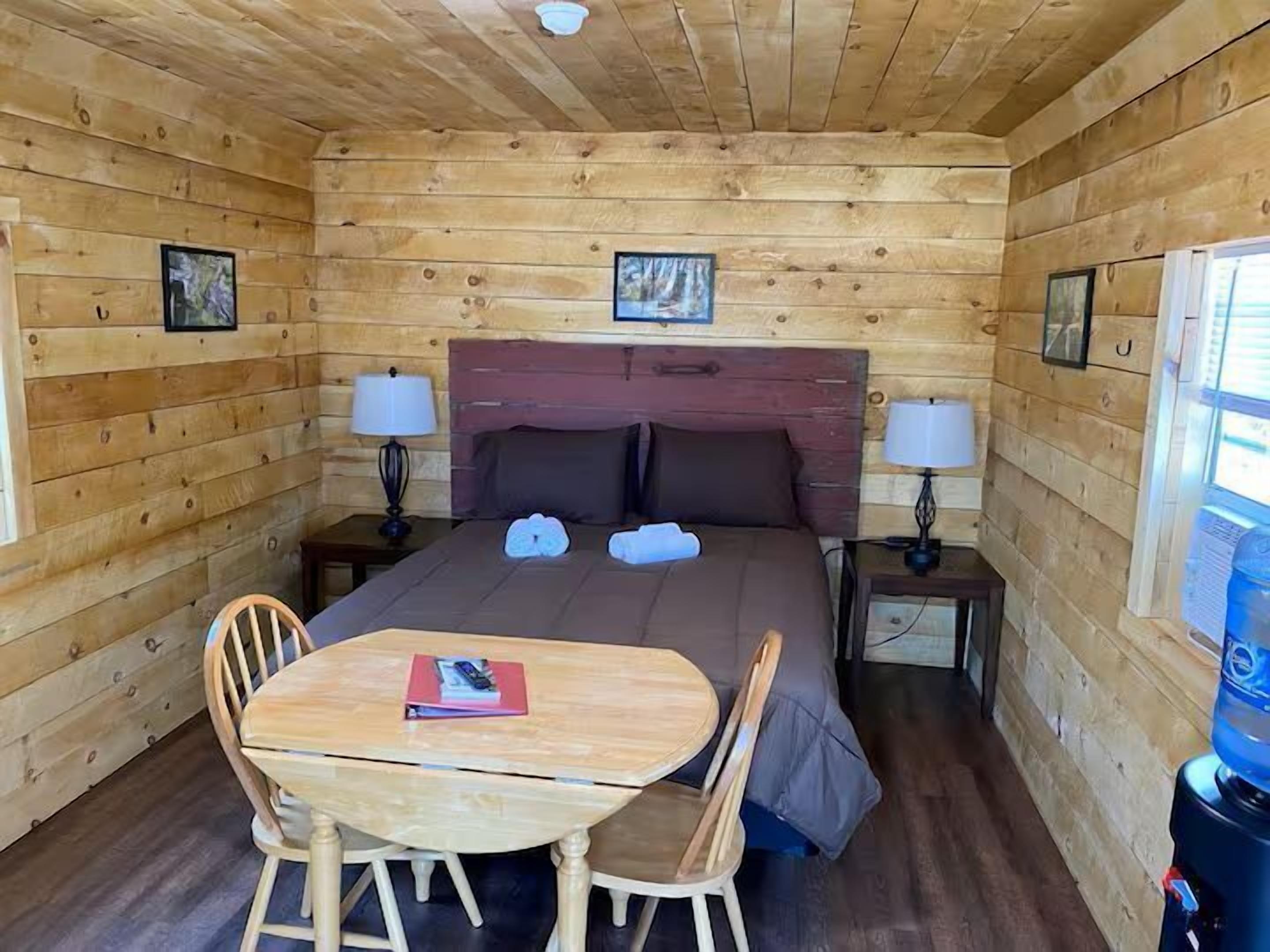 A wooden cabin room with a bed, two bedside tables, a table with chairs, and a window.