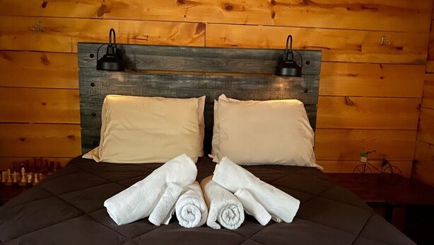 A bed with a dark quilted cover, two beige pillows, and a headboard with two wall-mounted lamps. - Canal Cabins (Paw Paw)