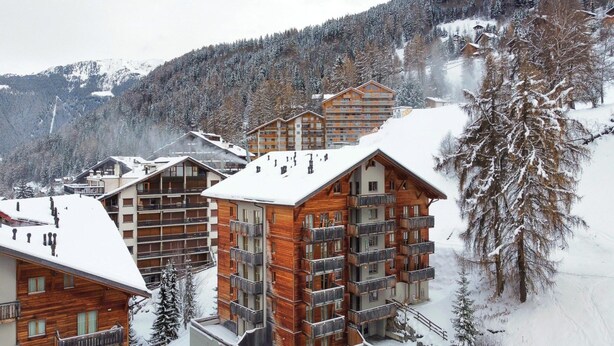 Una località turistica montana innevata con edifici in legno, balconi e una pista da sci sullo sfondo.