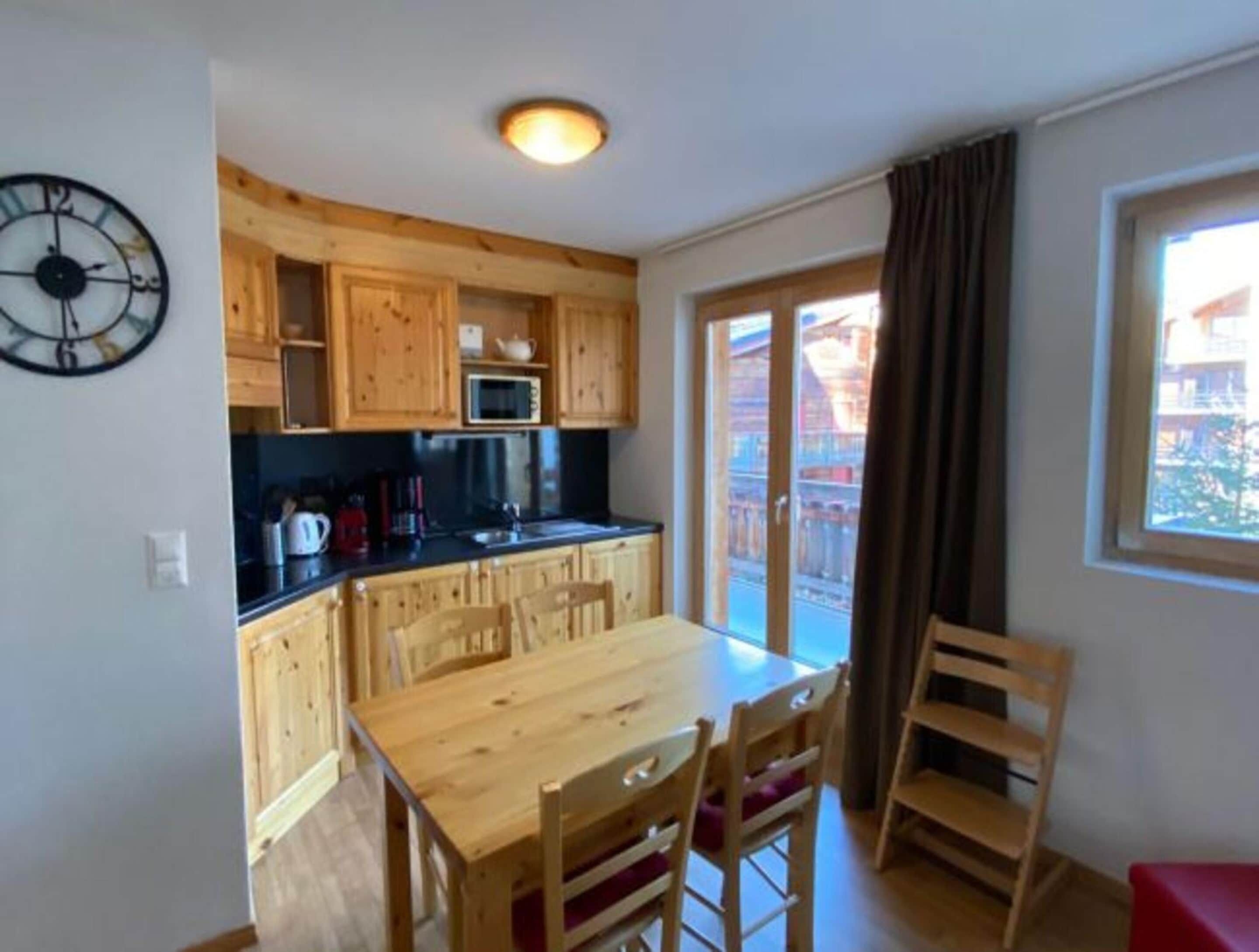 Cocina pequeña con armarios de madera, microondas y cafetera. Hay una mesa de comedor con sillas y una ventana con cortinas.