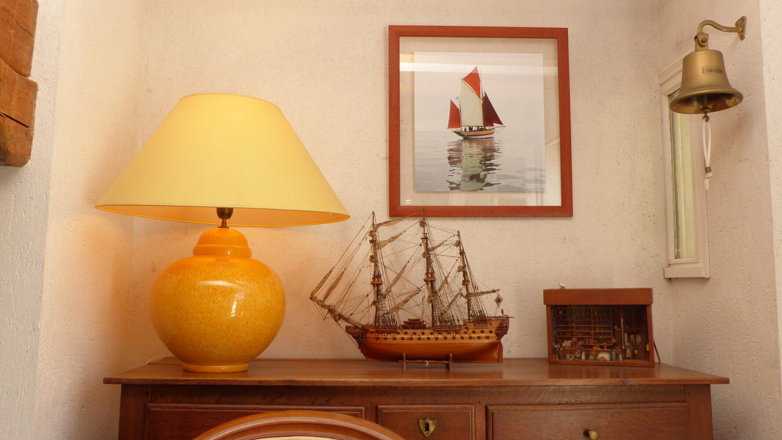 A wooden dresser with a yellow lamp, a framed picture of a sailboat, and a wall-mounted bell.