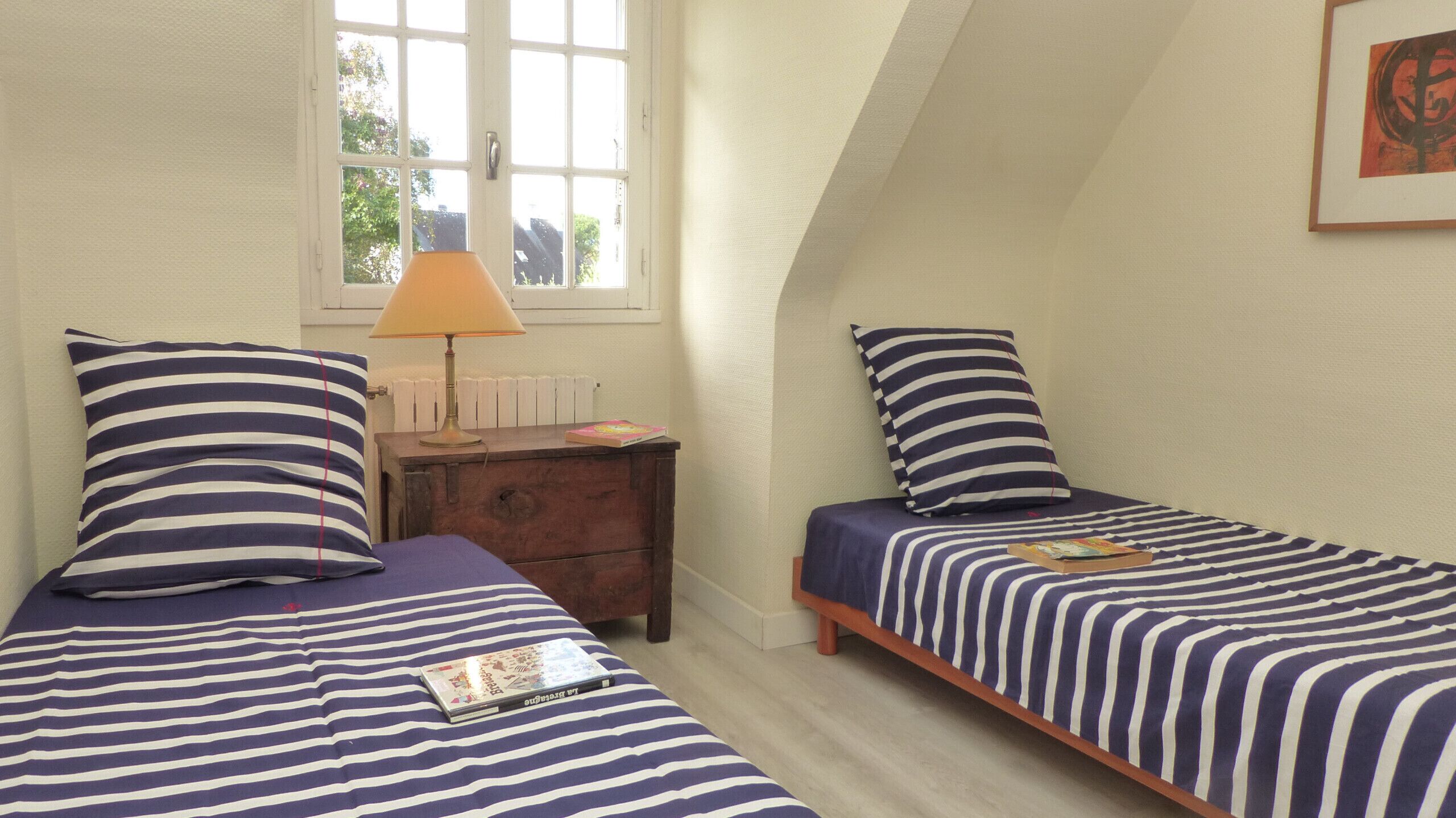 Un dormitorio con dos camas, cada una con ropa de cama a rayas, una mesita de noche con lámpara y una ventana con vistas a árboles.