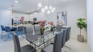 A modern kitchen with white cabinets, a central island, and a dining area with a glass table and chairs. - Gorgeous 5BR at Encore Resort next to Disney 7710 (Kissimmee)
