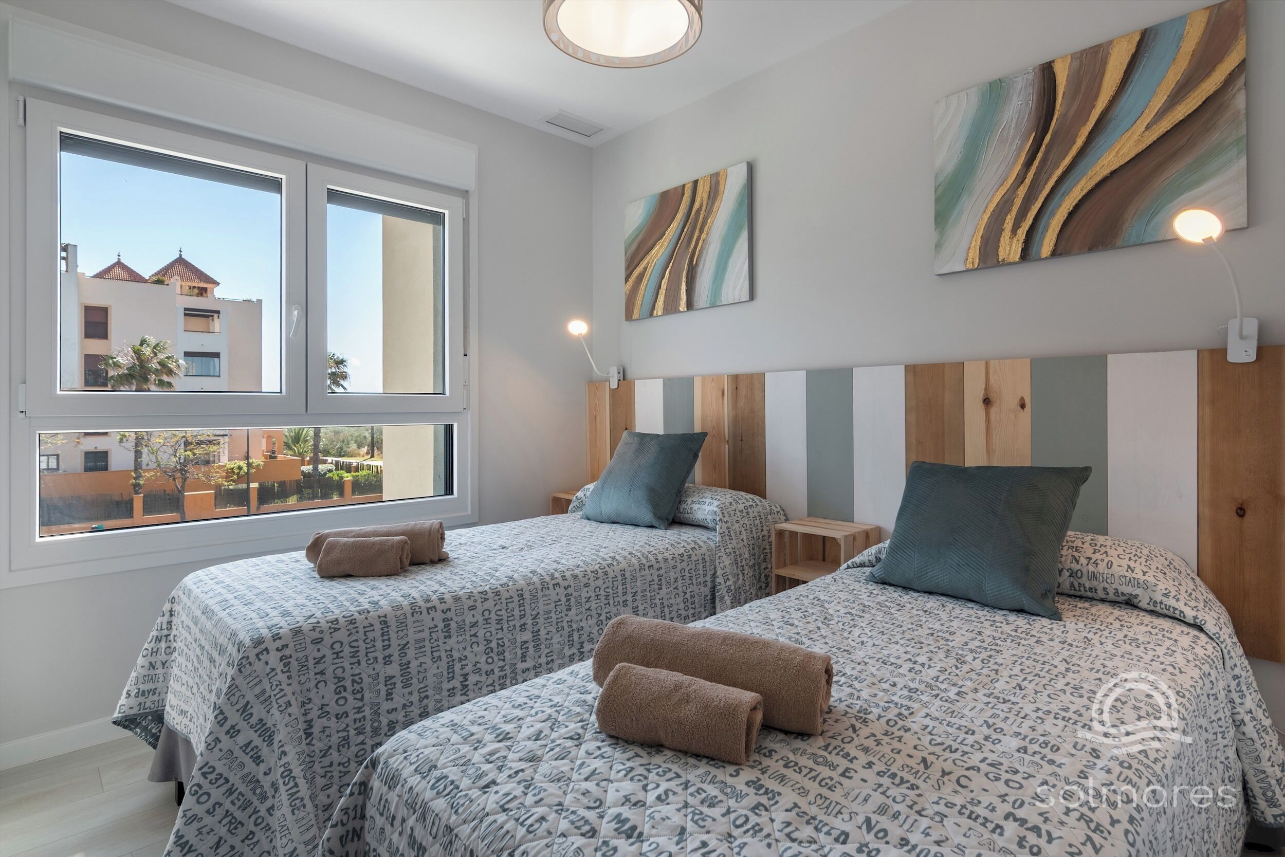 Ein Hotelzimmer mit zwei Betten, jeweils mit gemusterten Bettbezügen und Kissen, einem Holz-Kopfende, einem großen Fenster mit Blick auf Gebäude und einem an der Wand hängenden abstrakten Gemälde.