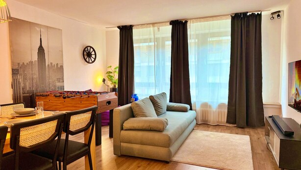 En opholdsstue med sofa, spisebord med stole, et fodboldbord og et stort vindue med gardiner.