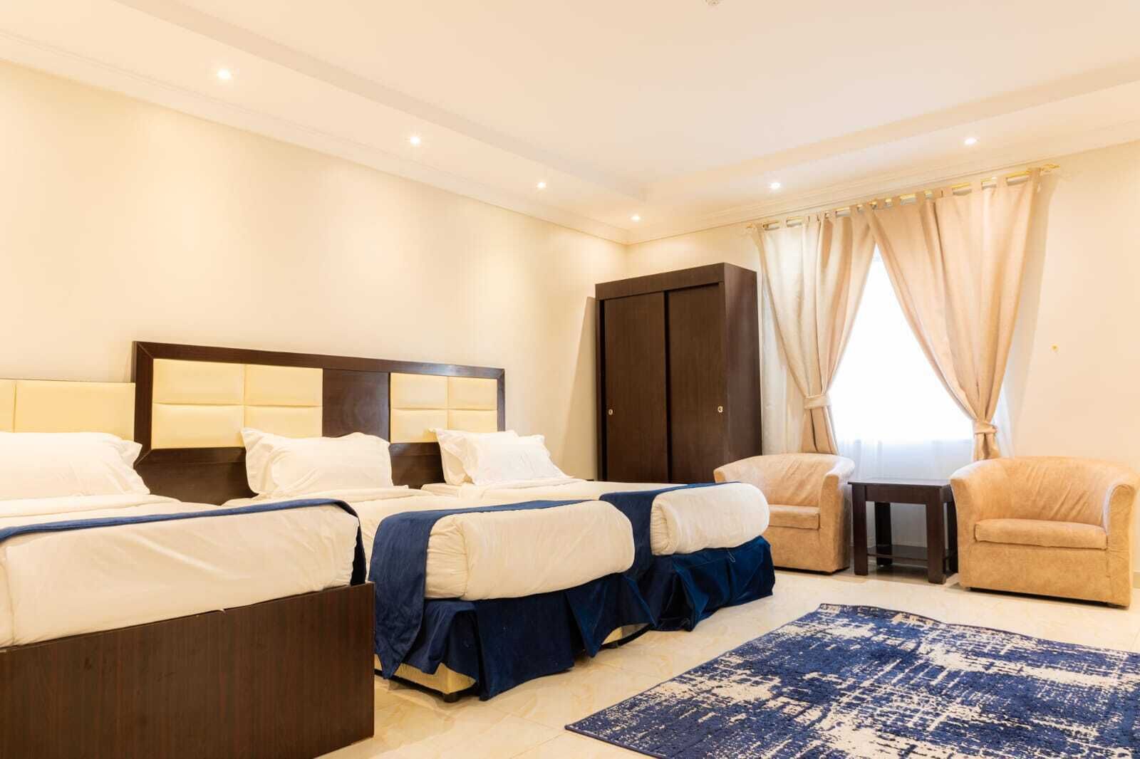 A hotel room with two beds, a wardrobe, a small table, two chairs, and a blue patterned rug.