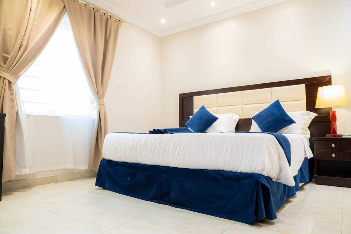 Habitación de hotel con una cama grande, una mesita de noche con lámpara y un ventanal con cortinas.