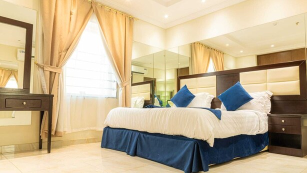 A well-lit bedroom with a large bed, a nightstand, a mirror, and a window with curtains. - Sweet Alnozha Serviced Apartments (Jeddah)